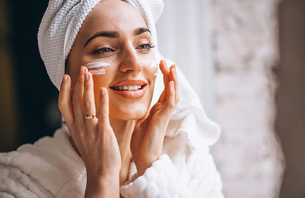 dra-debora-reis-procedimentos-bioestimulador-colageno-2 Mulher sorrindo enquanto aplica creme facial, simbolizando cuidados que estimulam o colágeno
